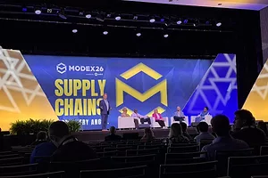 A man in a suit stands on a stage in front of four people sitting in chairs, in front of a large video screen that reads "SUPPLY CHAINS" 