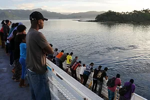 A group of people stand along the railing of a ferry off the coast of a lush, forested island