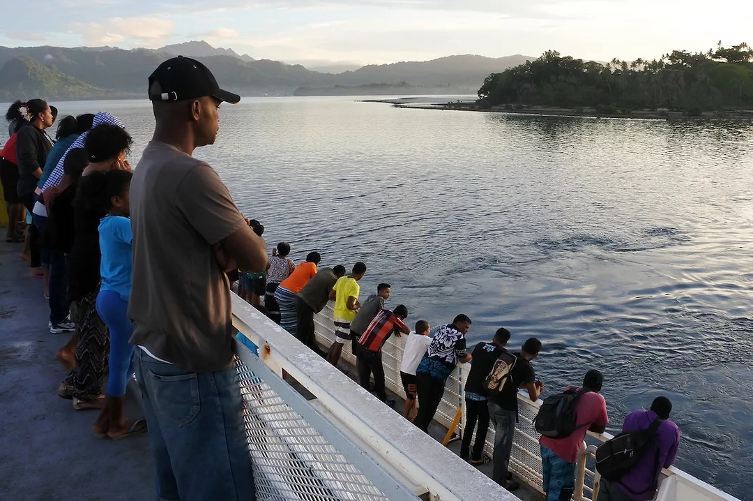 A group of people stand along the railing of a ferry off the coast of a lush, forested island