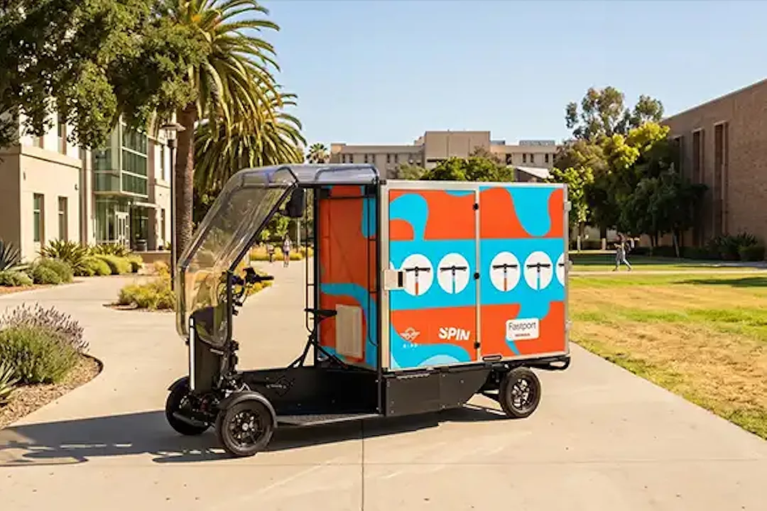 A small orange and blue box delivery vehicle sitting in a campus courtyard surrounded by tan buildings and greenery