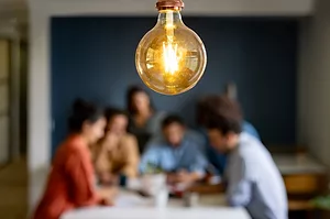 A LIGHTED LIGHT BULB HANGS OVER AN OUT OF FOCUS TABLE SURROUNDED BY PEOPLE DISCUSSING