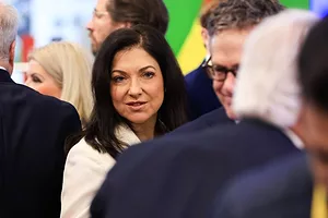 A WOMAN WITH LONG DARK HAIR IN A BEIGE JACKET IN A CROWD TURNS TO FACE THE CAMERA
