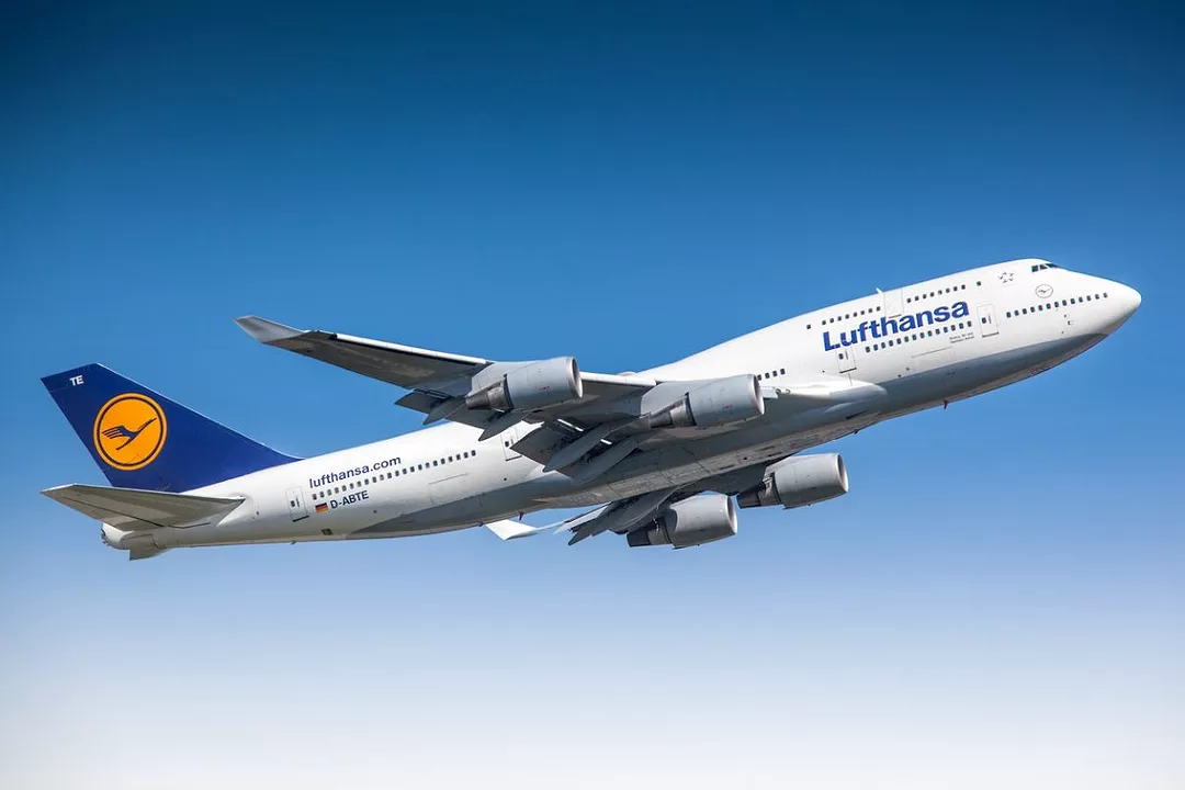 A PLANE BEARING THE LUFTHANSA BLUE AND GOLD INSIGNIA FLIES ACROSS A CLEAR BLUE SKY