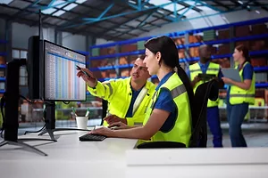 Two people in yellow safety vests sitting at a desk in a warehouse, with one pointing to a computer screen displaying a spreadsheet