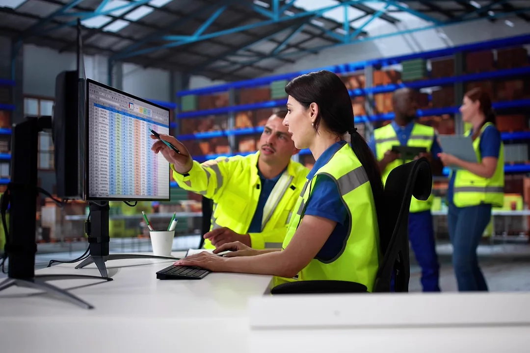 Two people in yellow safety vests sitting at a desk in a warehouse, with one pointing to a computer screen displaying a spreadsheet