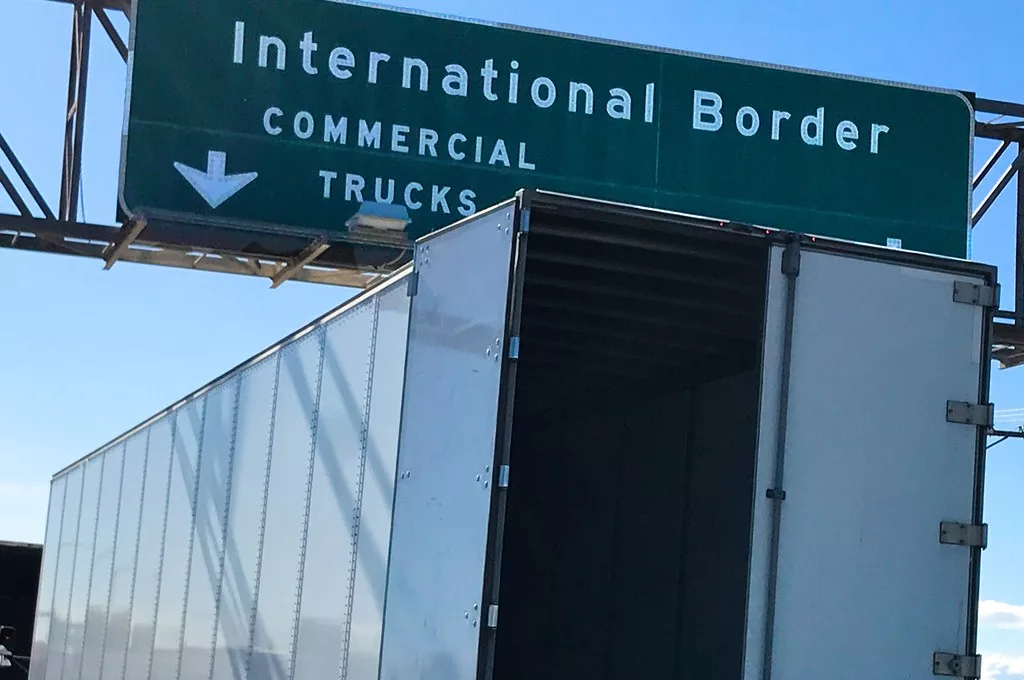 A TRUCK WITH ITS CONTAINER DOOR OPEN SITS UNDER A SIGN THAT READS INTERNATIONAL BORDER COMMERCIAL TRUCKS