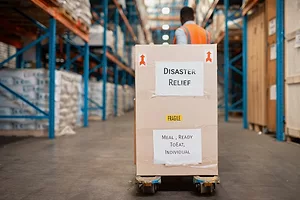 a man in an orange safety vest in a warehouse pulling a handcart with a large box on it that reads "Disaster Relief"