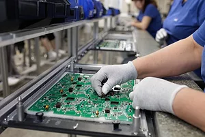A green circuit board being assembled by a worker on a factory assembly line