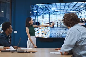 A woman in a green dress in a conference room gesturing to a screen picturing a warehouse, while three employees sit the conference table looking on