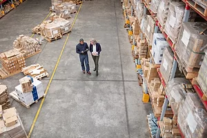 Two men in suits walking down the aisle of a warehouse stacked with shelves holding pallets and boxes