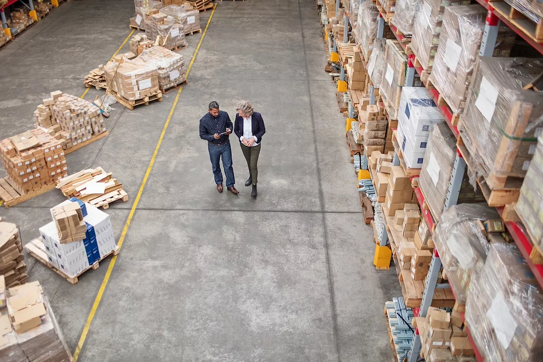 Two men in suits walking down the aisle of a warehouse stacked with shelves holding pallets and boxes