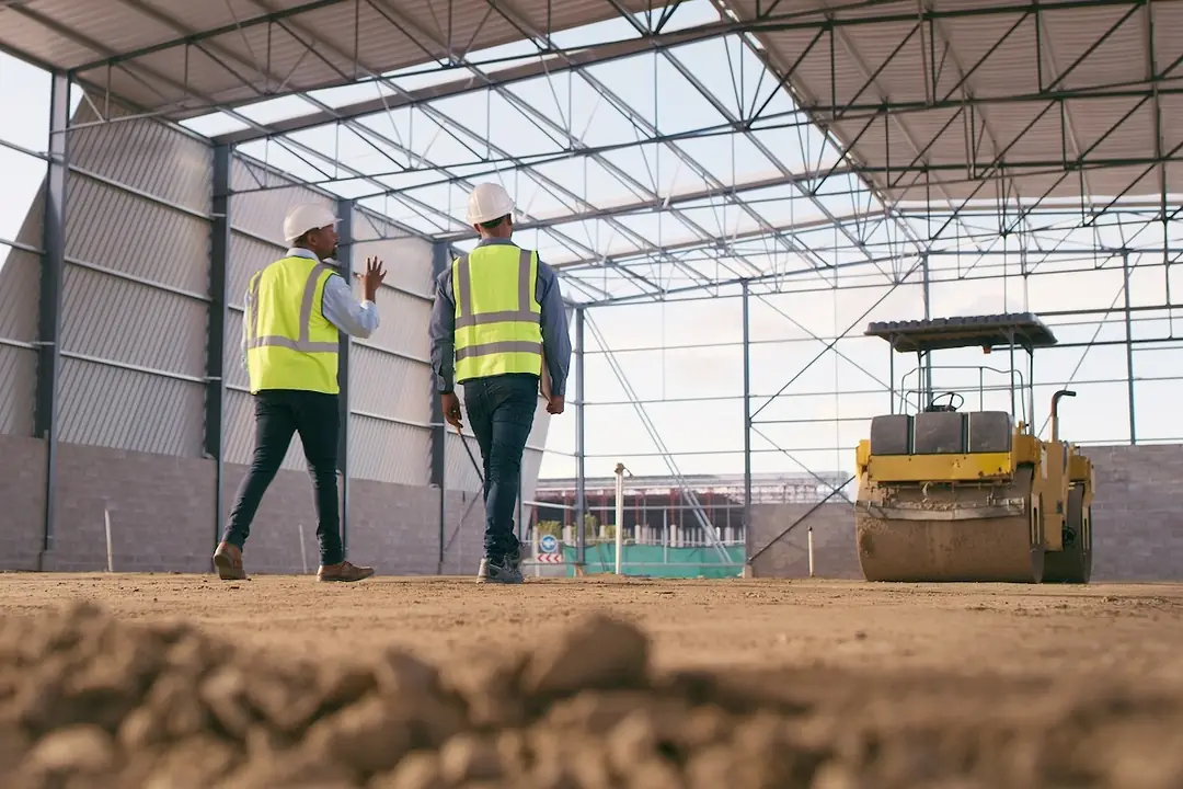 Two men in yellow safety vests and helmets walking under a half-built warehouse facility