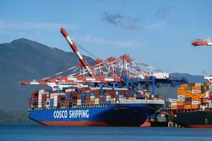 A COSCO CONTAINER SHIP SITS AT DOCK UNDER RED AND WHITE STRIPED CRANES