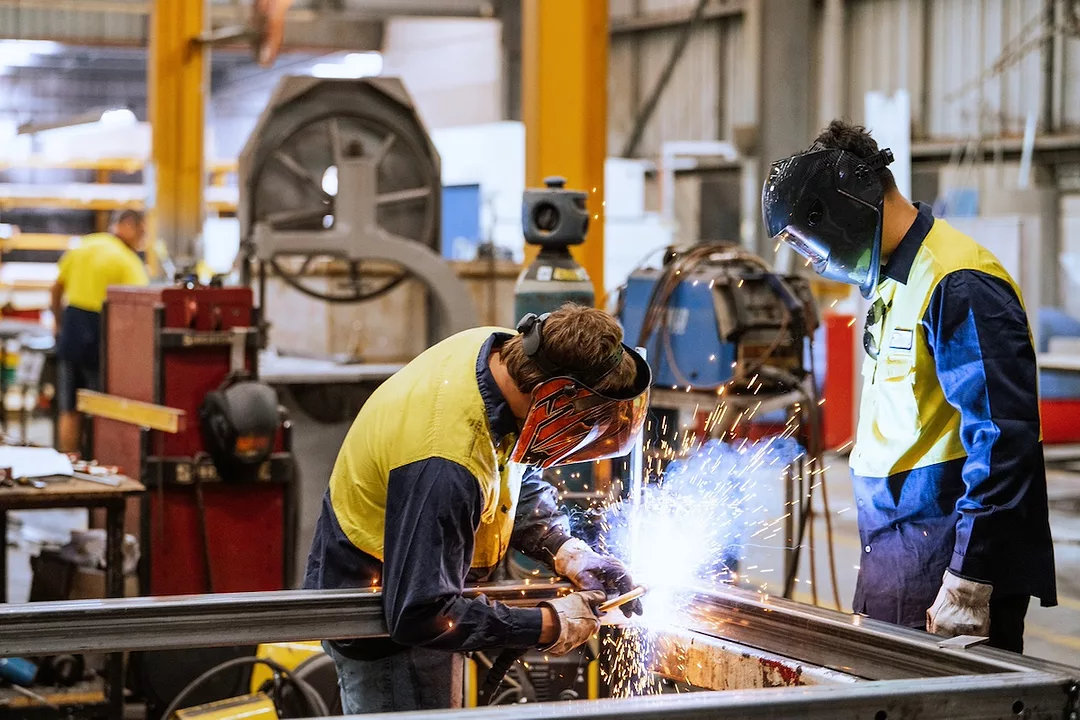 Two men in welding masks in a factory, with the one of the left using a welding torch on a square metal frame.
