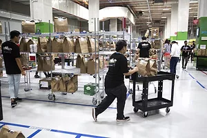 A man in a black tshirt and jeans pushing a cart full of brown grocery bags, next to a rack lined with similar bags