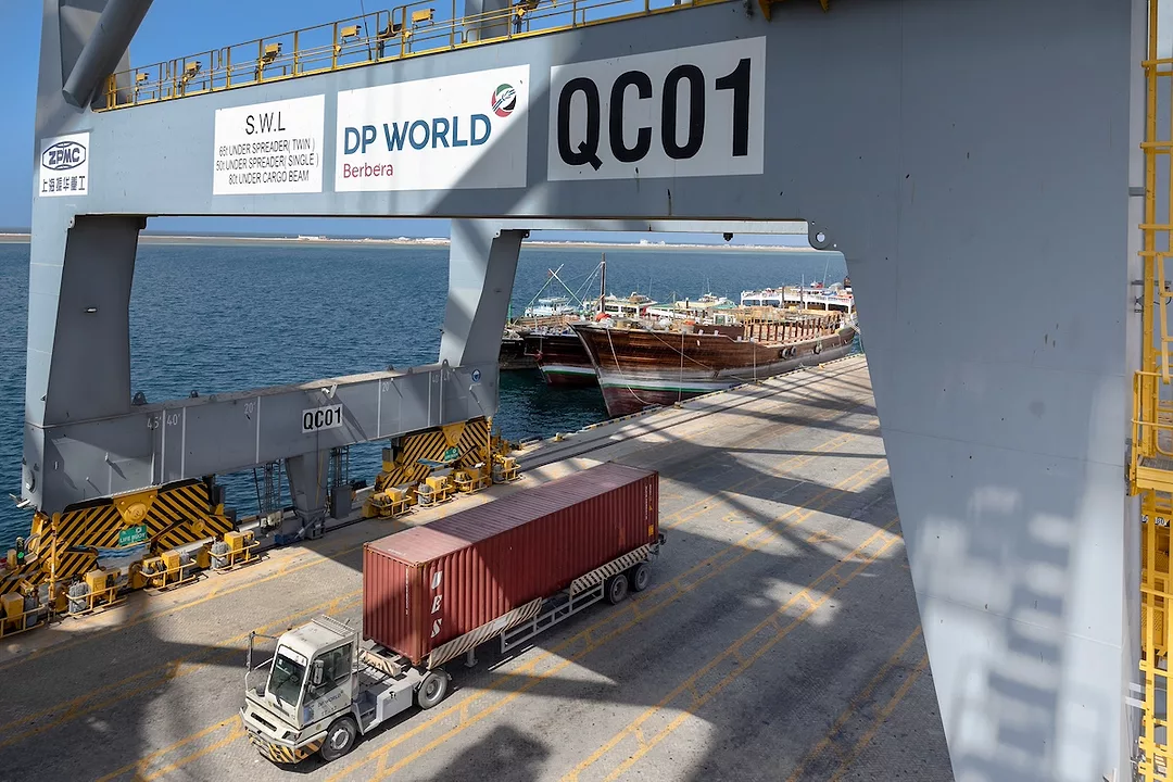 A semi-truck pulling a red shipping container drives underneath a crane along a shoreline