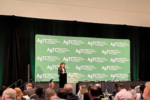 A woman in a suit speaking on stage, in front of a green banner that reads "AgTC"
