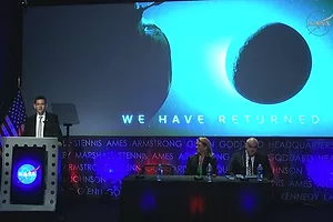 A man in a suit standing at a NASA-emblazoned podium, in front of a screen depicting the moon, which reads "We have returned"