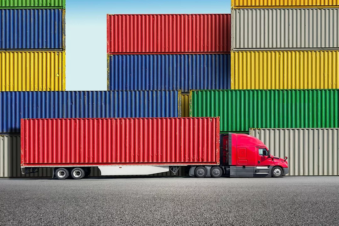 A red semi-truck towing a red container in front of a stack of multi-colored shipping containers