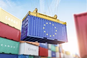 A blue shipping container with an image of the EU flag on it, being held by a crane above the ground and surrounded by other multi-colored containers 