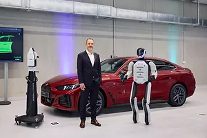 A SMILING MAN IN A SUIT STANDS NEXT TO A HUMANOID ROBOT IN FRONT OF A RED CAR