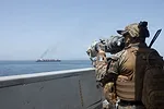 A FIGURE IN CAMOUFLAGE LOOKS THROUGH A SCOPING DEVICE AT A SHIP IN THE DISTANCE, BELCHING SMOKE