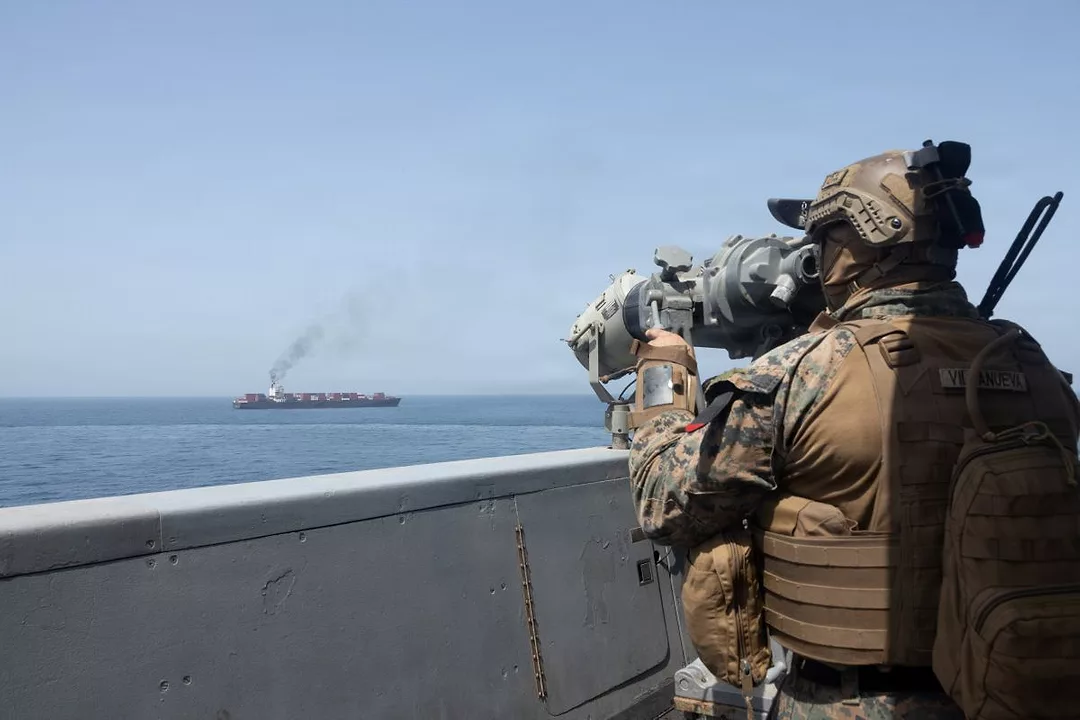 A FIGURE IN CAMOUFLAGE LOOKS THROUGH A SCOPING DEVICE AT A SHIP IN THE DISTANCE, BELCHING SMOKE
