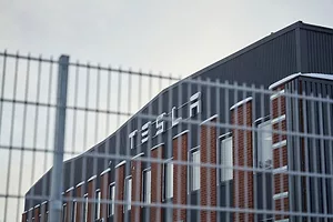 A LARGE BLACK AND BROWN FACTORY BEARING THE TESLA LOGO IS SEEN THROUGH A CHAIN LINK FENCE