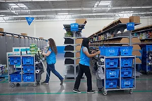 TWO WORKERS IN A WAREHOUSE PUSH ROLLING CARTS LOADED WITH BRIGHT BLUE BINS