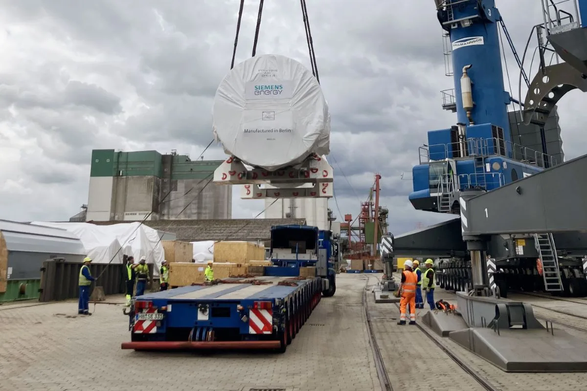 A LARGE CYLINDRICAL OBJECT SHRINK-WRAPPED IN WHITE PLASTIC IS LOWERED BY CRANE ONTO A FLAT BED TRUCK ON A DOCK