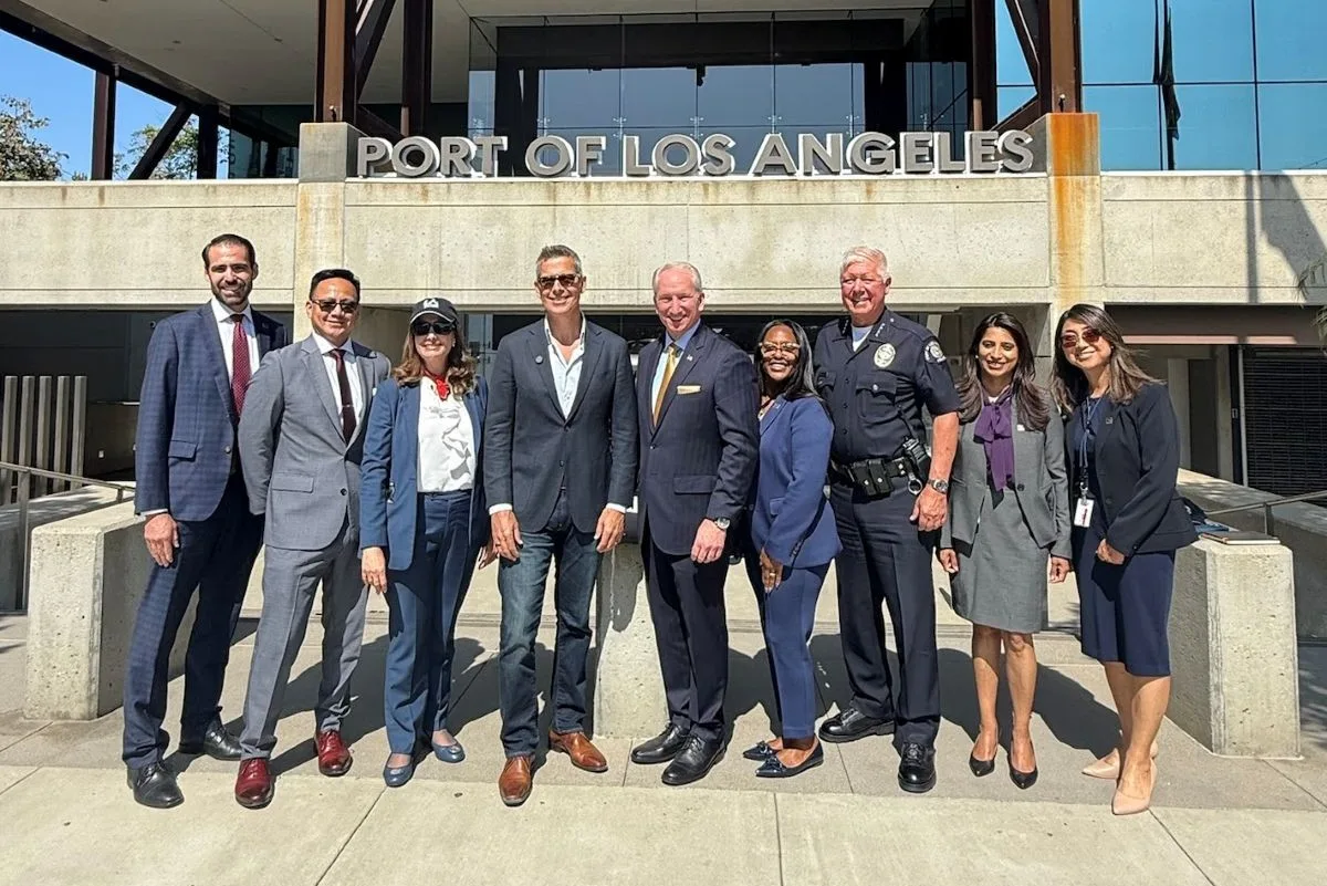 A GROUP OF NINE PEOPLE STAND SMILING IN A ROW IN THE SUNSHINE BENEATH A SIGN SAYING PORT OF LOS ANGELES