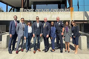 A GROUP OF NINE PEOPLE STAND SMILING IN A ROW IN THE SUNSHINE BENEATH A SIGN SAYING PORT OF LOS ANGELES