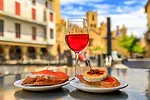 A GLASS OF ROSE WINE IS FLANKED BY PLATES OF FOOD, A MEDIEVAL TOWN SQUARE OUT OF FOCUS IN THE BACKGROUND