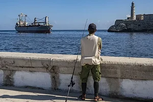 A MAN WITH A FISHING ROD LOOKS OUT AT A SHIP SAILING PAST A LIGHTHOUSE
