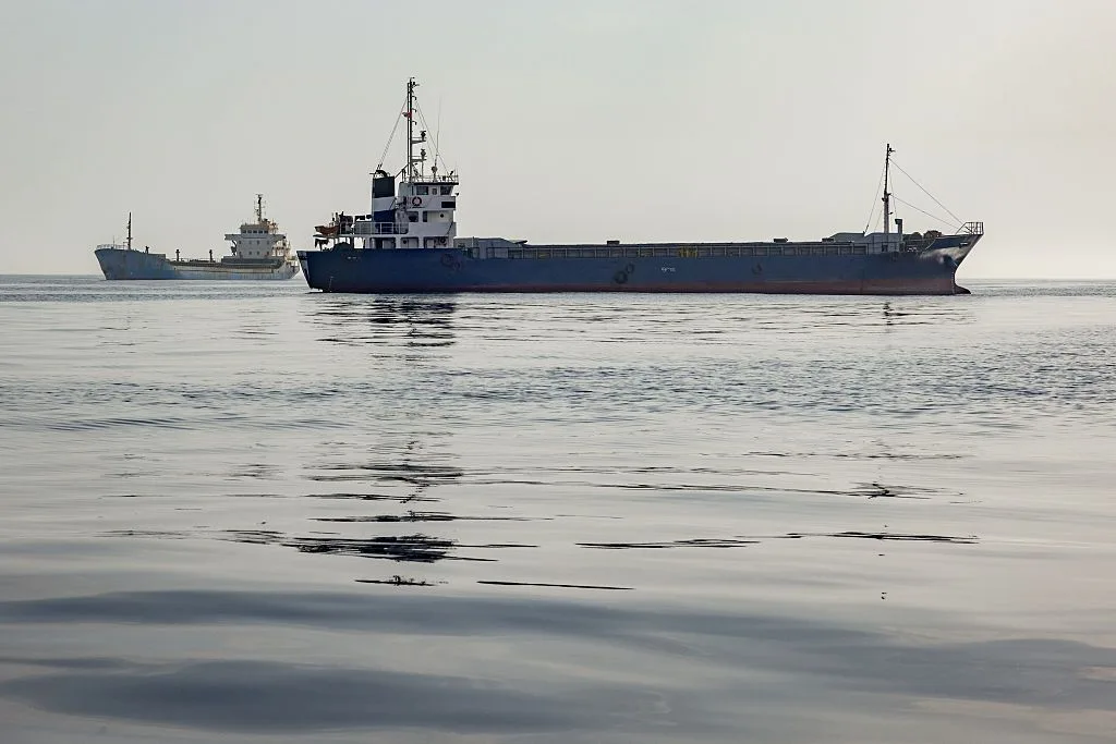 A SHIP SITS ON CALM SEA, ANOTHER SHIP VISIBLE BEHIND IT