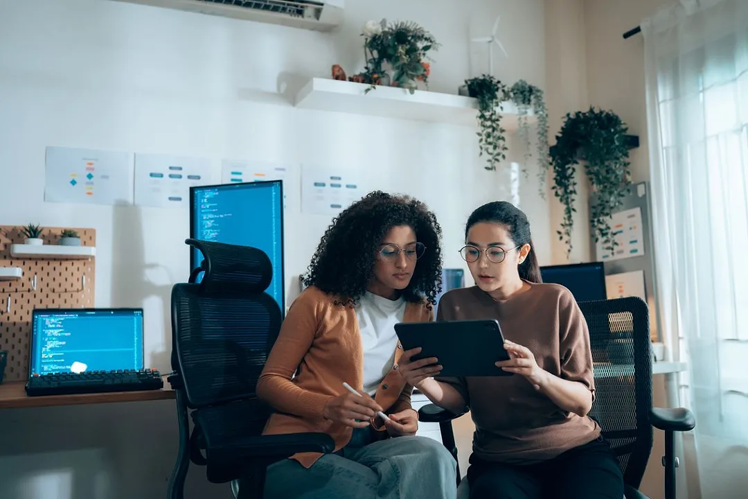 Two Women collaborate over a tablet with coding monitors in the background