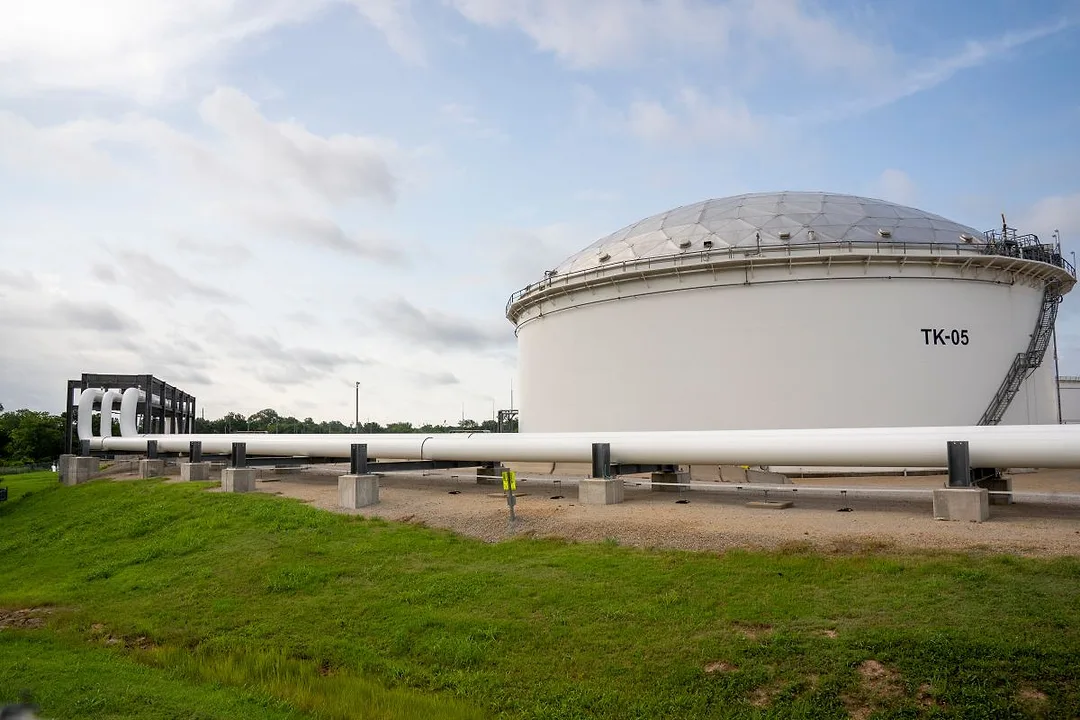 A LARGE WHITE OIL STORAGE TANK CROUCHES IN A FIELD, A PIPELINE RUNNING FROM IT