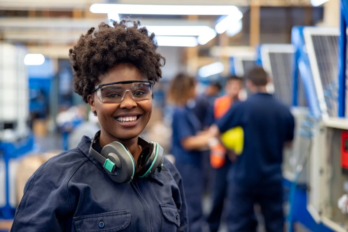 A SMILING WORKER IN BLUE OVERALLS AND EAR PROTECTIVE HEADPHONES STANDS IN A FACTORY