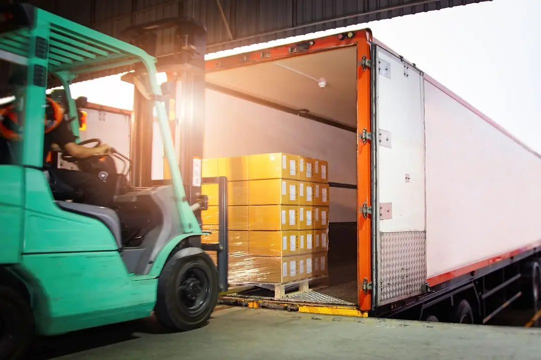 A WORKER OPERATES A FORKLIFT TO UNLOAD PALLETS FROM A TRUCK IN A LOADING DOCK
