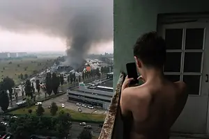 A CHILD STANDS ON A BALCONY, FILMING A RISING PLUME OF SMOKE IN THE DISTANCE WITH A SMARTPHONE