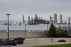 BEHIND HIGH WALLS IN A PARKING LOT, INDUSTRIAL STACKS RISE