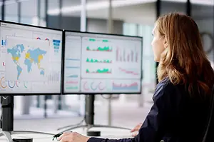 A Business Woman Looks At two computer screens showing data Dashboards