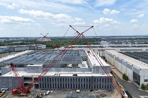 A white warehouse building under construction, with red cranes towering over the top