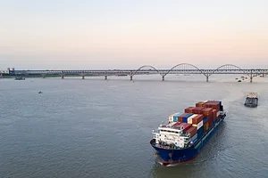 A blue container ship moving toward a long bridge in the distance
