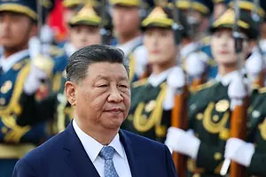 A MAN IN A BLUE SUIT AND RED TIE STANDS SCOWLING IN FRONT OF RANKS OF MEN IN MILITARY UNIFORMS