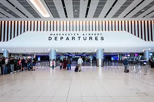 An airport departures terminal, with "Manchester Airport" written in black lettering on a white overhang, as travelers walk through the terminal