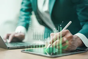 A PERSON IN A BUSINESS SUIT LEANS OVER A LAPTOP, TYPING, AN IMAGE OF A GRAPH SUPER-IMPOSED ON TOP