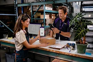 A WORKER IN AN APRON HANDS A PACKAGE TO A CUSTOMER OVER A COUNTER, BOTH WIELDING CELLPHONES