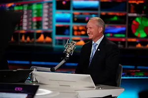 A MAN IN A SUIT STANDS SPEAKING INTO A MICROPHONE IN A TV STUDIO, GLOWING SCREENS IN THE BACKGROUND