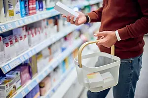 A man in a red sweater holding a small box, while standing in the pharmacy aisle of a store